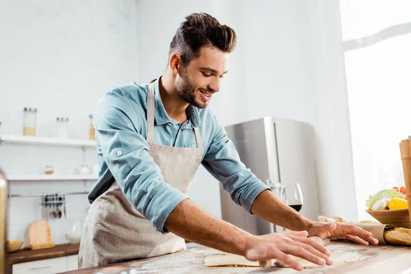 Jovem sorridente em avental com rolo de pino preparando massa na cozinha — Fotografia de Stock