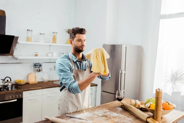 Schöner junger Mann in Schürze mit ungekochtem Teig bei der Zubereitung von Pizza — Stockfoto