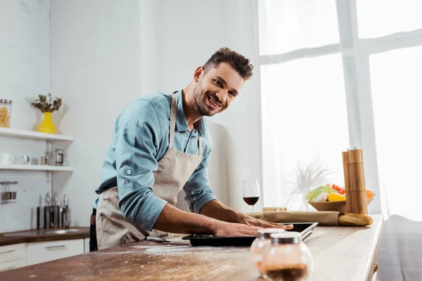 Handsome young man in apron smiling at camera while preparing pizza dough on baking tray — Stock Photo