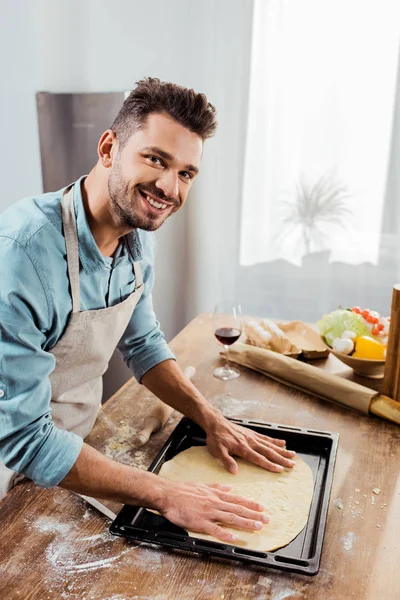 Giovane sorridente in grembiule preparare la pasta della pizza sulla teglia — Foto stock