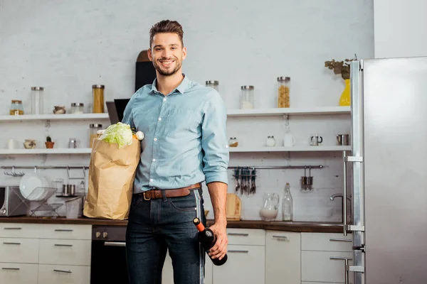 Joven guapo con bolsa de comestibles y botella de vino sonriendo a la cámara en la cocina - foto de stock