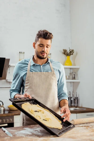 Frustré jeune homme dans tablier regardant la pâte gâtée sur plaque de cuisson — Photo de stock