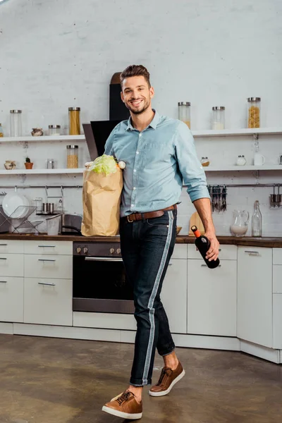 Felice bell'uomo con borsa della spesa e bottiglia di vino sorridente alla macchina fotografica in cucina — Foto stock