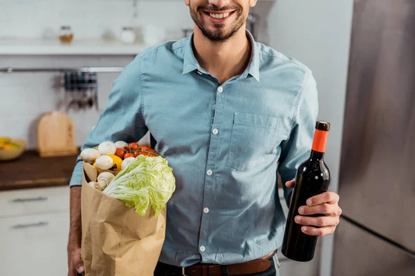 Recortado disparo de sonriente joven sosteniendo bolsa de comestibles y botella de vino en la cocina - foto de stock