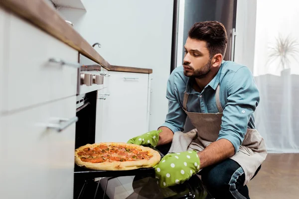 Young man in apron and potholders taking out baking tray with pizza from oven — Stock Photo