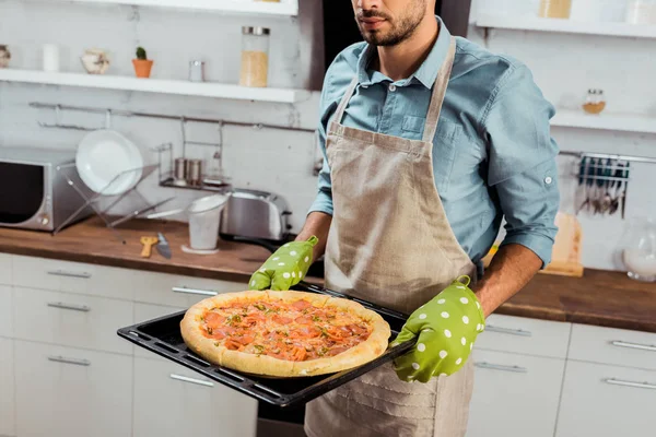 Tiro recortado de hombre en delantal y agarraderas sosteniendo bandeja para hornear con pizza casera fresca - foto de stock