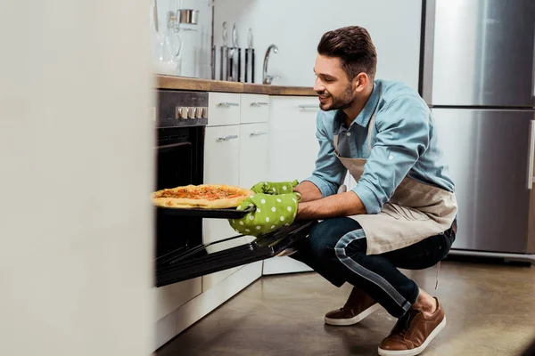 Smiling young man in apron taking out baking tray with pizza from oven — Stock Photo