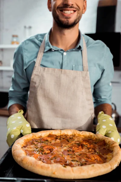 Cropped shot of smiling man in apron and potholders holding baking tray with fresh homemade pizza — Stock Photo