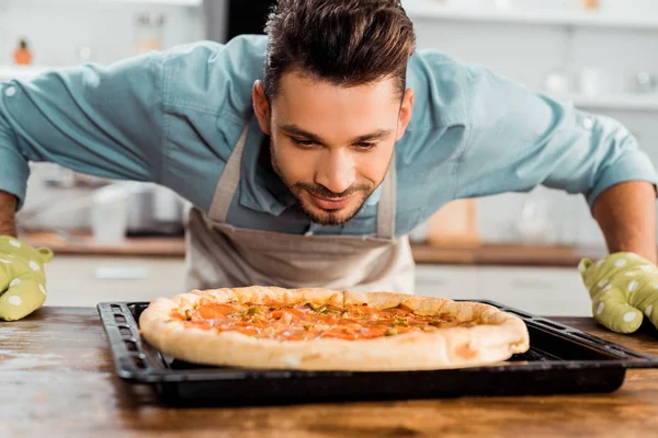 Guapo sonriente joven oliendo pizza fresca casera en bandeja para hornear - foto de stock