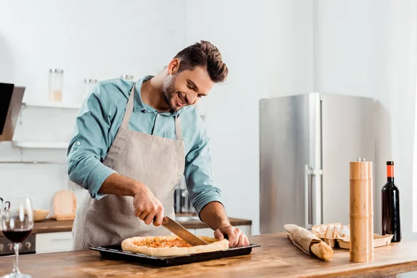 Счастливый молодой человек в фартуке режет свежую домашнюю пиццу на подносе для выпечки — стоковое фото