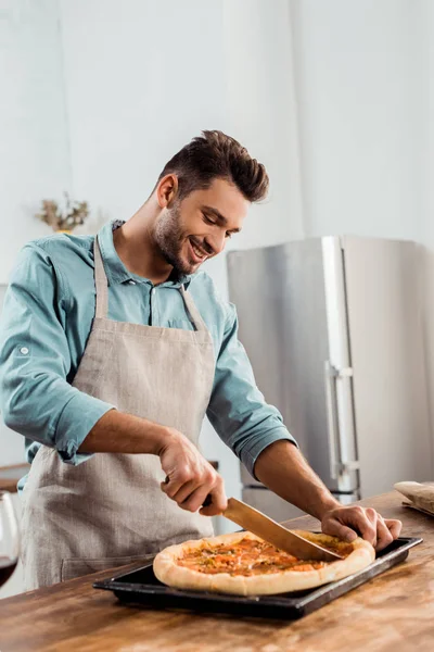 Jeune homme souriant dans tablier coupe pizza fraîche maison sur plaque de cuisson — Photo de stock