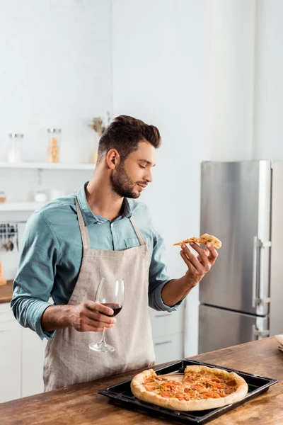 Jovem bonito em avental segurando fatia de pizza caseira e copo de vinho — Fotografia de Stock