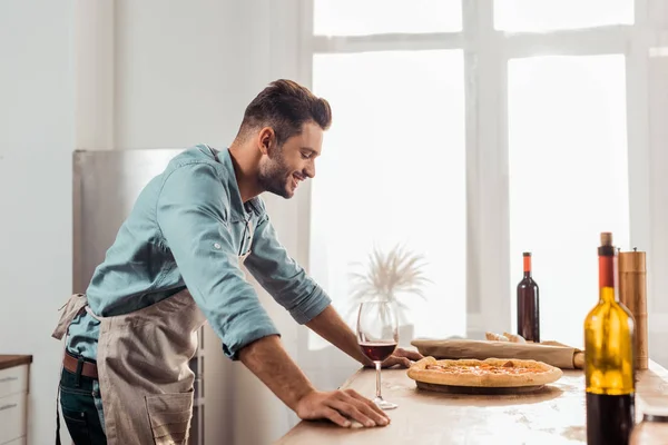 Vista lateral del joven sonriente en aron mirando pizza casera fresca - foto de stock