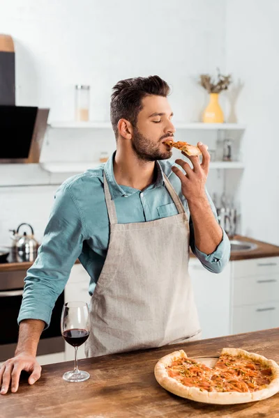 Giovane in grembiule mangiare pizza fatta in casa in cucina — Foto stock