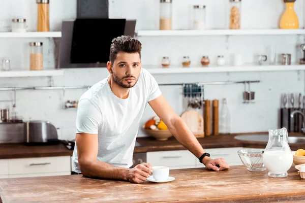 Молодий чоловік тримає чашку кави і дивиться на камеру на кухні вранці — стокове фото