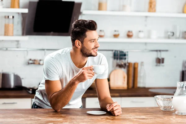 Sorrindo jovem segurando xícara de café e inclinando-se na mesa da cozinha de manhã — Fotografia de Stock