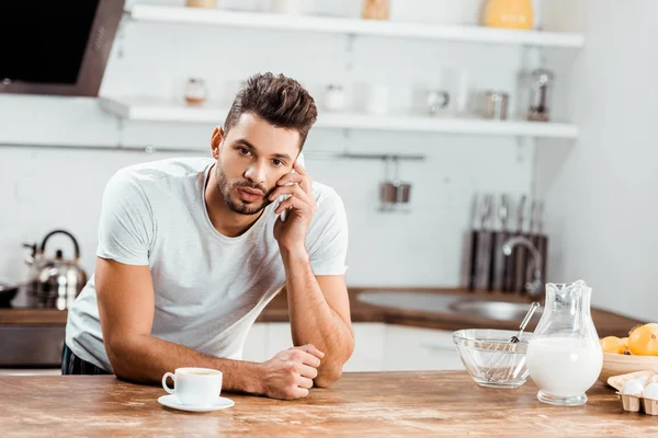 Молодой человек склоняется за кухонным столом и разговаривает со смартфоном по утрам — стоковое фото