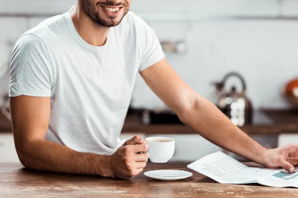 Recortado disparo de sonriente joven sosteniendo taza de café y periódico por la mañana - foto de stock