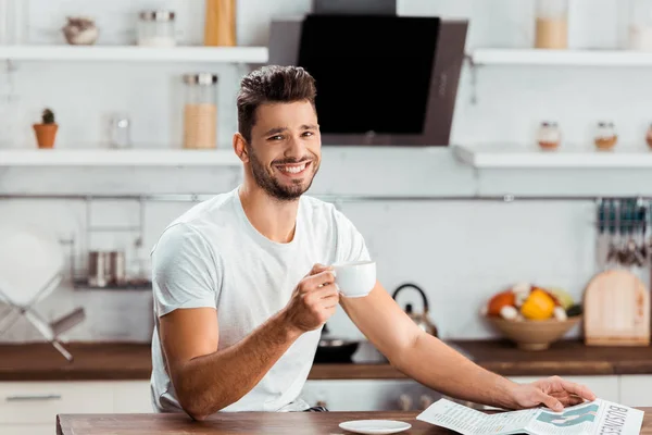 Handsome young man holding cup of coffee and smiling at camera in kitchen — Stock Photo