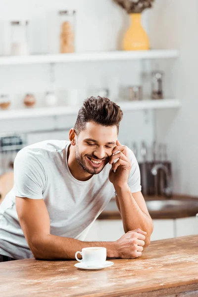 Giovane uomo sorridente che parla con lo smartphone mentre si appoggia al tavolo della cucina con una tazza di caffè — Foto stock