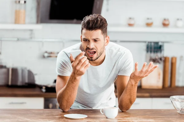Emotional young man yelling at smartphone while drinking coffee in kitchen — Stock Photo