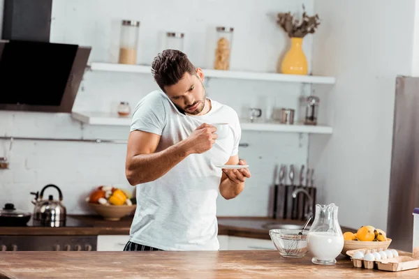 Young man holding cup of coffee and talking by smartphone in kitchen — Stock Photo