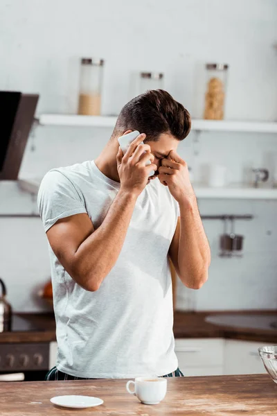 Frustrated young man rubbing nose bridge and talking by smartphone at morning in kitchen — Stock Photo