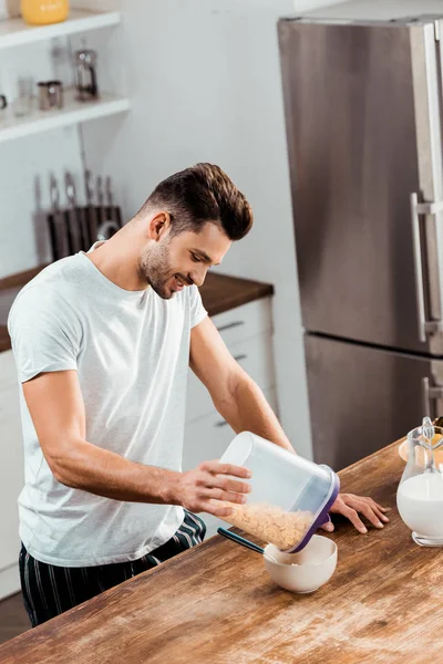 Visão de alto ângulo do jovem sorridente segurando recipiente com flocos de milho e preparando café da manhã — Fotografia de Stock