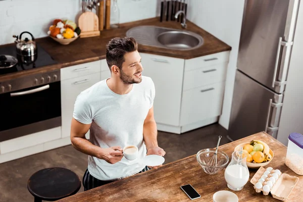 Vista de ángulo alto del joven sonriente sosteniendo la taza de café y mirando hacia otro lado en la cocina - foto de stock