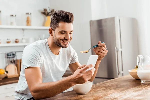 Giovane sorridente utilizzando lo smartphone e facendo colazione a casa — Foto stock