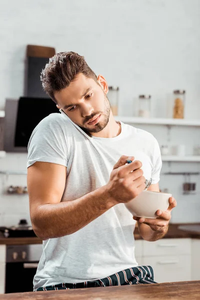 Young man in pajamas eating breakfast and talking by smartphone — Stock Photo