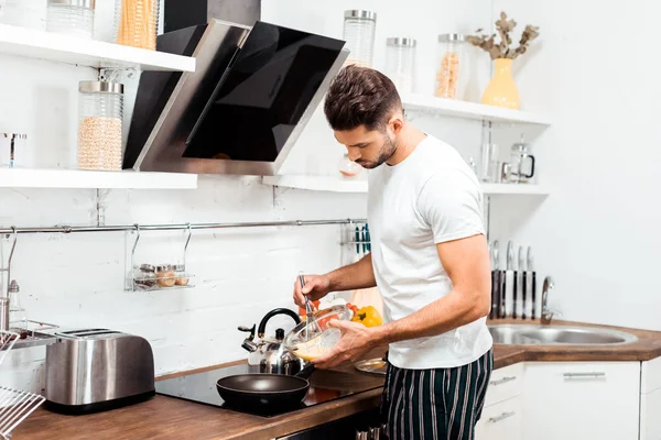 Красивий молодий чоловік у піжамі готує омлет на сковороді вранці — стокове фото