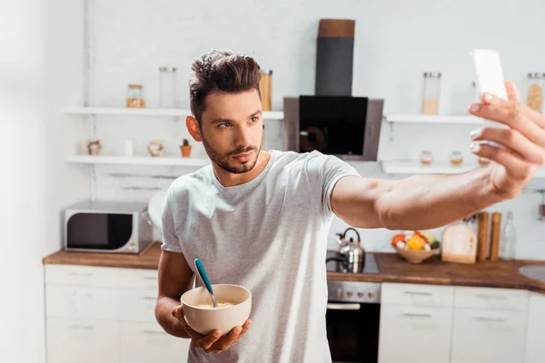 Bel giovanotto che tiene la ciotola con la colazione e prende selfie con smartphone in cucina — Foto stock