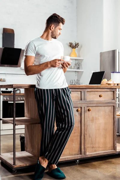 Joven en pijama sosteniendo una taza de café y mirando el portátil en la cocina - foto de stock