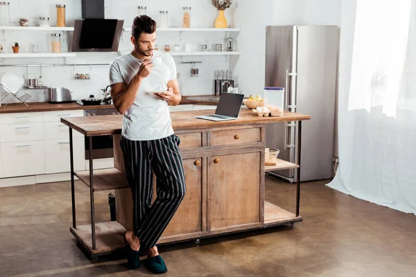 Bell'uomo giovane in pigiama che beve caffè in cucina — Foto stock
