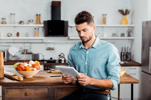 Красивый молодой человек, использующий цифровой планшет во время приготовления пищи на кухне — стоковое фото