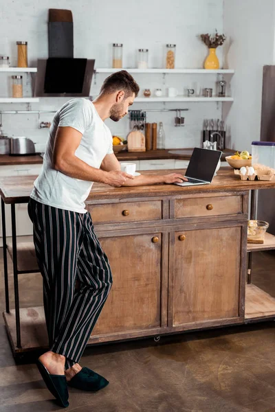 Jovem em pijama usando laptop e segurando xícara de café na cozinha — Fotografia de Stock