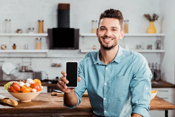 Bel giovanotto che tiene lo smartphone con lo schermo vuoto e sorride alla fotocamera in cucina — Foto stock