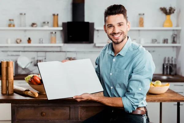 Handsome young man holding blank cookbook and smiling at camera in kitchen — Stock Photo