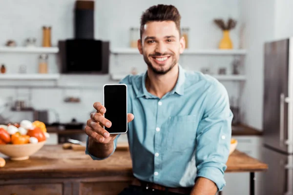 Joven guapo sosteniendo teléfono inteligente con pantalla en blanco y sonriendo a la cámara en la cocina - foto de stock