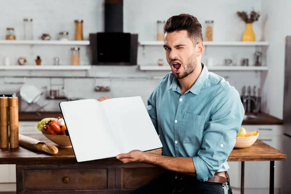 Schöner junger Mann hält leeres Kochbuch in der Küche und zwinkert in die Kamera — Stockfoto