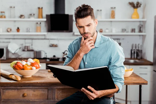 Thoughtful handsome young man reading cookbook in kitchen — Stock Photo