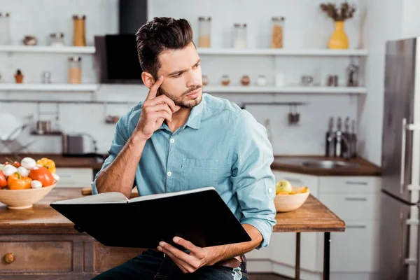 Pensive young man holding cookbook and looking away in kitchen — Stock Photo