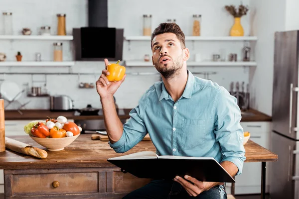 Young man holding fresh pepper and cookbook while sitting in kitchen and looking up — Stock Photo