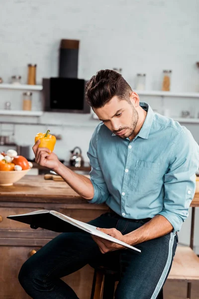 Focused young man holding fresh pepper and reading cookbook in kitchen — Stock Photo