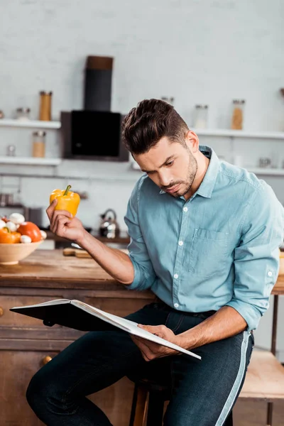 Focado jovem segurando pimenta fresca e leitura de livro de receitas na cozinha — Fotografia de Stock