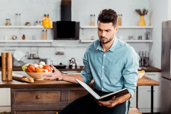Handsome young man throwing fresh pepper and reading cookbook in kitchen — Stock Photo