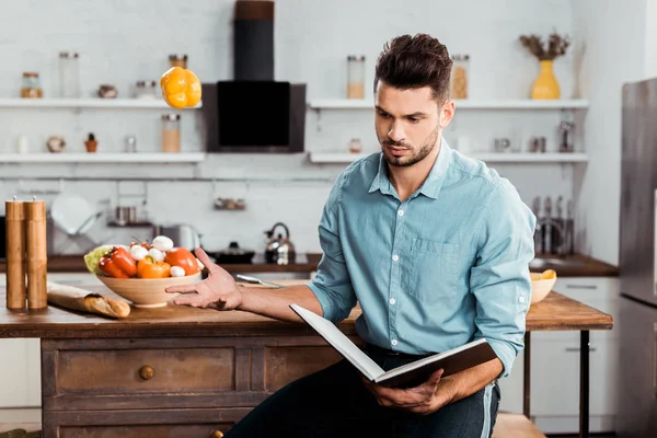 Joven guapo lanzando pimienta fresca y leyendo libro de cocina en la cocina - foto de stock