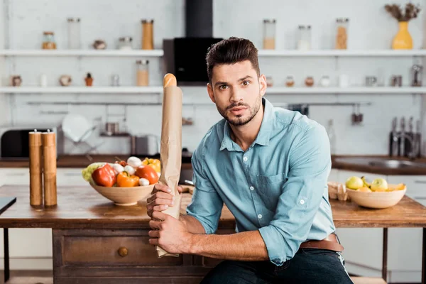 Schöner junger Mann mit frischem Baguette in Papiertüte und Blick in die Kamera in der Küche — Stockfoto