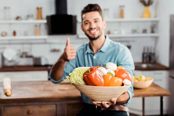 Sonriente joven sosteniendo tazón con verduras frescas y mostrando el pulgar hacia arriba en la cocina - foto de stock
